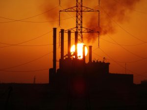 A dramatic silhouette of a power plant with smoke stacks and power lines against a golden sunset.