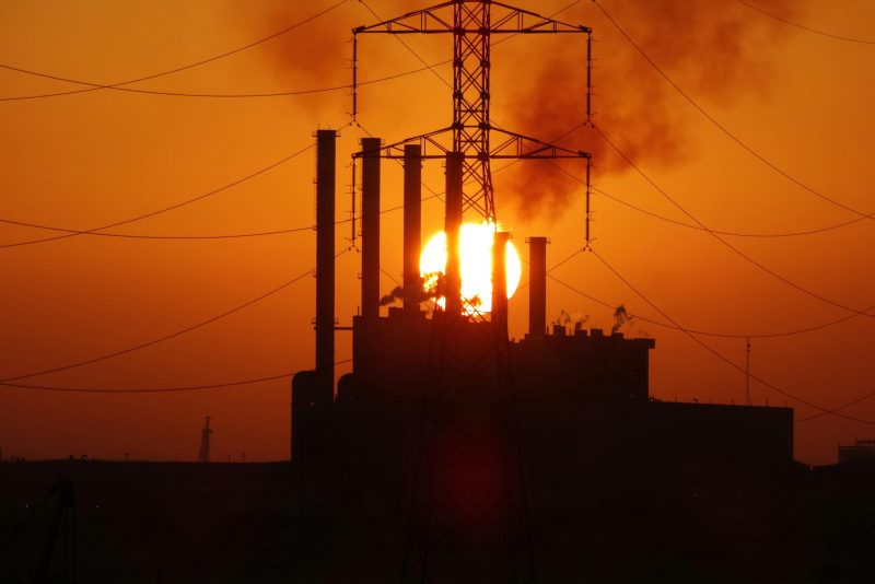 A dramatic silhouette of a power plant with smoke stacks and power lines against a golden sunset.